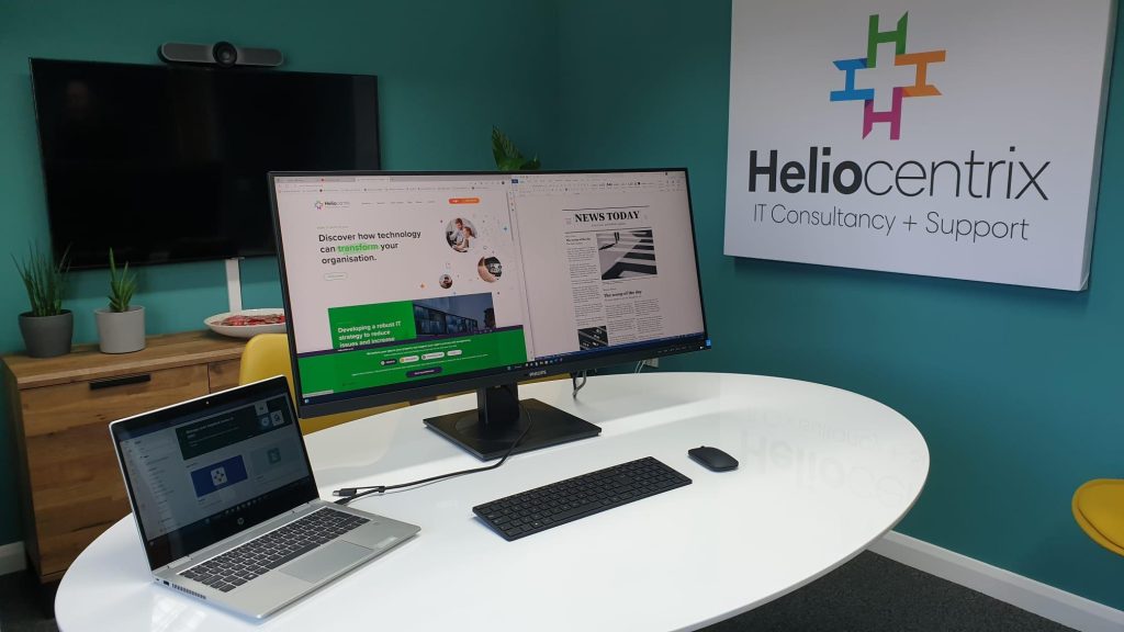 A HP X360 laptop docked with a Philips Ultrawide monitor with the Heliocentrix company logo in the background