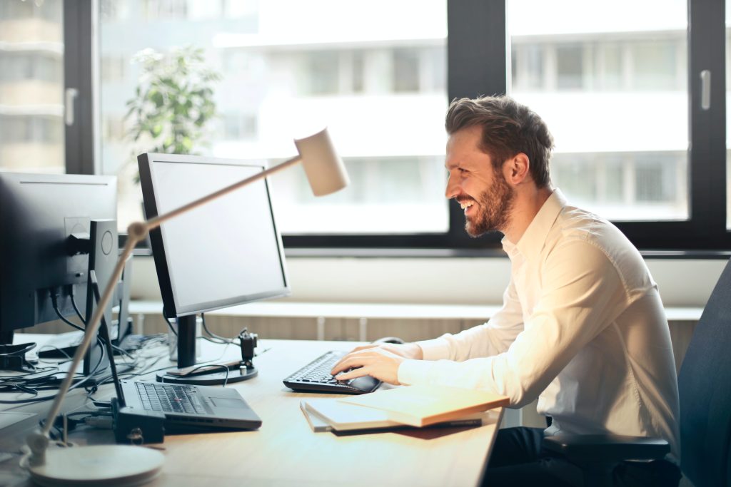 A man sitting at a desk looking at a computer monitor and smiling. 