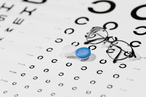 A pair of glasses and contact lenses placed on a opticians eye testing board where the letters get smaller and smaller as you look down the board. 