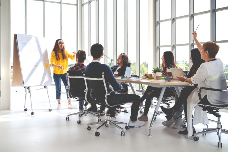 Modern office with employees working collaboratively around a big desk, contributing to a flipchart at the front of the room