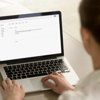 Businesswoman typing e-mail on laptop at office desk, composing professional email letter using business etiquette, writing e message to corporate client online, focus on screen, close up rear view