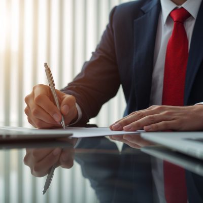 Image is a professional, high-resolution photograph featuring a business setting. The layout is focused on a close-up view of a person’s hands and upper body, dressed in a dark navy suit with a white shirt and a red tie. The individual is using a silver laptop, positioned on a reflective surface, possibly a desk. The person is holding a pen in their right hand, poised over a piece of paper, suggesting they are taking notes or signing a document. The background is softly blurred, with vertical blinds partially open, allowing natural light to filter through, creating a warm ambiance. The overall composition conveys a sense of professionalism and focus.