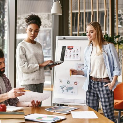 Attractive young woman standing near whiteboard with a laptop in her hands and looking to her male colleague, that holds a chart with the results. Another colleague looks at it with a smile. Concept of success