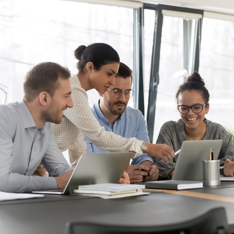 Smiling diverse young colleagues gather at desk in office look at laptop screen brainstorming at meeting together, happy multiracial employees cooperate using computer at briefing, teamwork concept