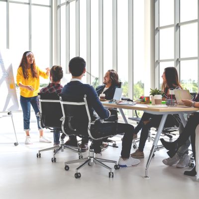 Modern office with employees working collaboratively around a big desk, contributing to a flipchart at the front of the room
