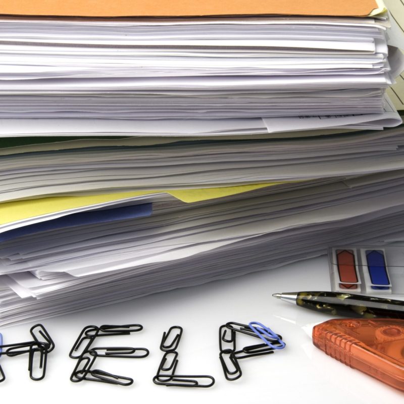 Lots of paperwork on a desk with ‘HELP’ spelled in paper clips